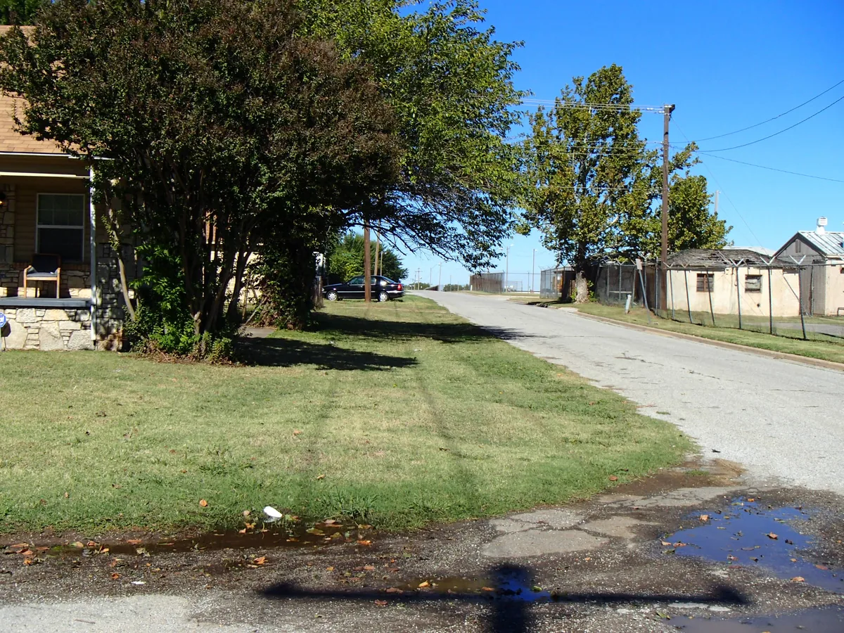 A grassy side yard beside a stone house, with a street on the right and fenced bus-lot buildings across it, in daylight with the trees in full leaf