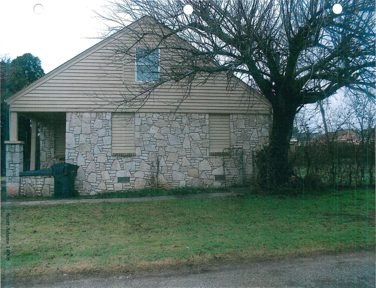 Side of a stone house with covered windows, a bare tree and a strip of grass along the street